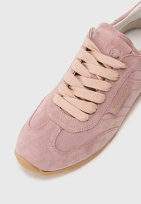 Baskets en daim rose avec des lacets beiges, présentant des coutures courbées et une semelle en caoutchouc. Détail de marque doré sur le côté.