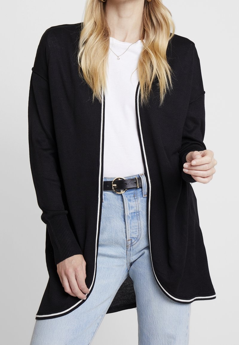 Černý pletený kardigan s pohodlným střihem, bílým lemem po okrajích a žebrovanými rukávy. Kombinován s bílým tričkem a světle modrými džínami.