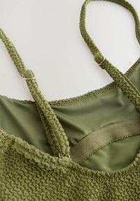 Maslinasto zeleni bikini gornji deo sa texturiranim, nabranim materijalom. Podesive naramenice imaju zlatne detalje. Glatka unutrašnja podstava upotpunjuje dizajn.