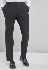 Pantalons noirs bien ajustés, avec une texture lisse, des poches latérales et une fermeture à bouton. Portés avec des chaussures en cuir noires.