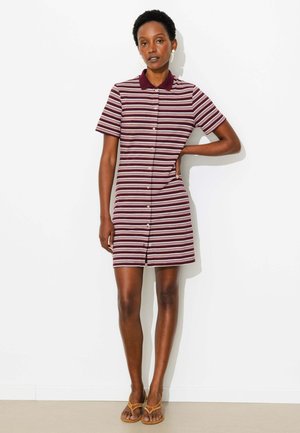 Femme portant une robe boutonnée à manches courtes, rayée bordeaux et blanche avec un col bordeaux et des sandales beiges, debout avec une main sur la hanche.