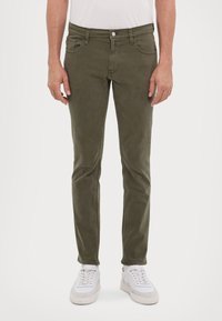 Olíva zöld, slim-fit farmernadrág, pamutból készült, elülső zsebekkel, cipzáras állással és egygombos záródással, fehér sneakers cipővel párosítva.
