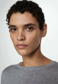 Krupan plan osobe s kratkom kovrčavom kosom koja nosi sivi džemper. Osobine uključuju glatku kožu i suptilne pjegice, uz neutralni make-up.