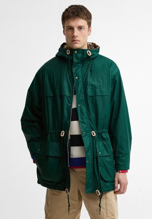 Mees tumerohelises kapuutsiga jopes, mitmevärvilise triibulise kampsuni ja beežide pükstega, seisab ühevärvilise valge tausta ees.