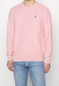 Pull en tricot rose clair à manches longues, avec poignets et ourlet côtelés ; présente un petit logo bleu sur la poitrine ; texture douce.