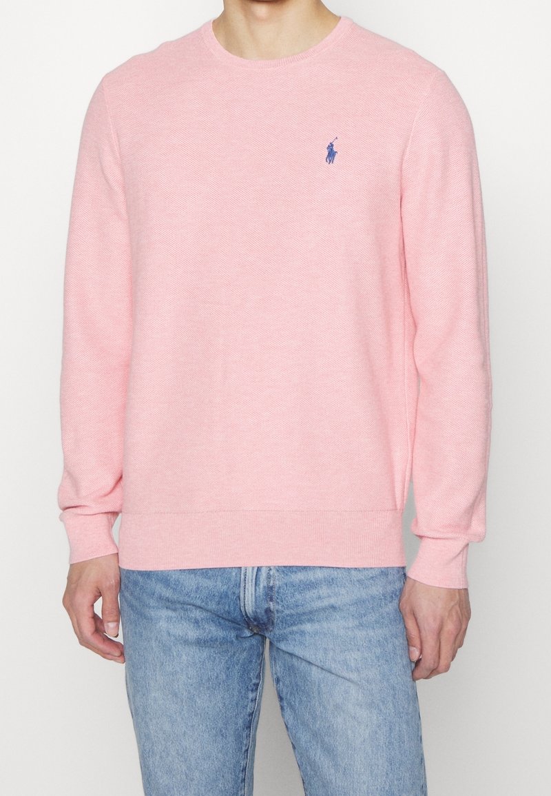 Pull en tricot rose clair à manches longues, avec poignets et ourlet côtelés ; présente un petit logo bleu sur la poitrine ; texture douce.