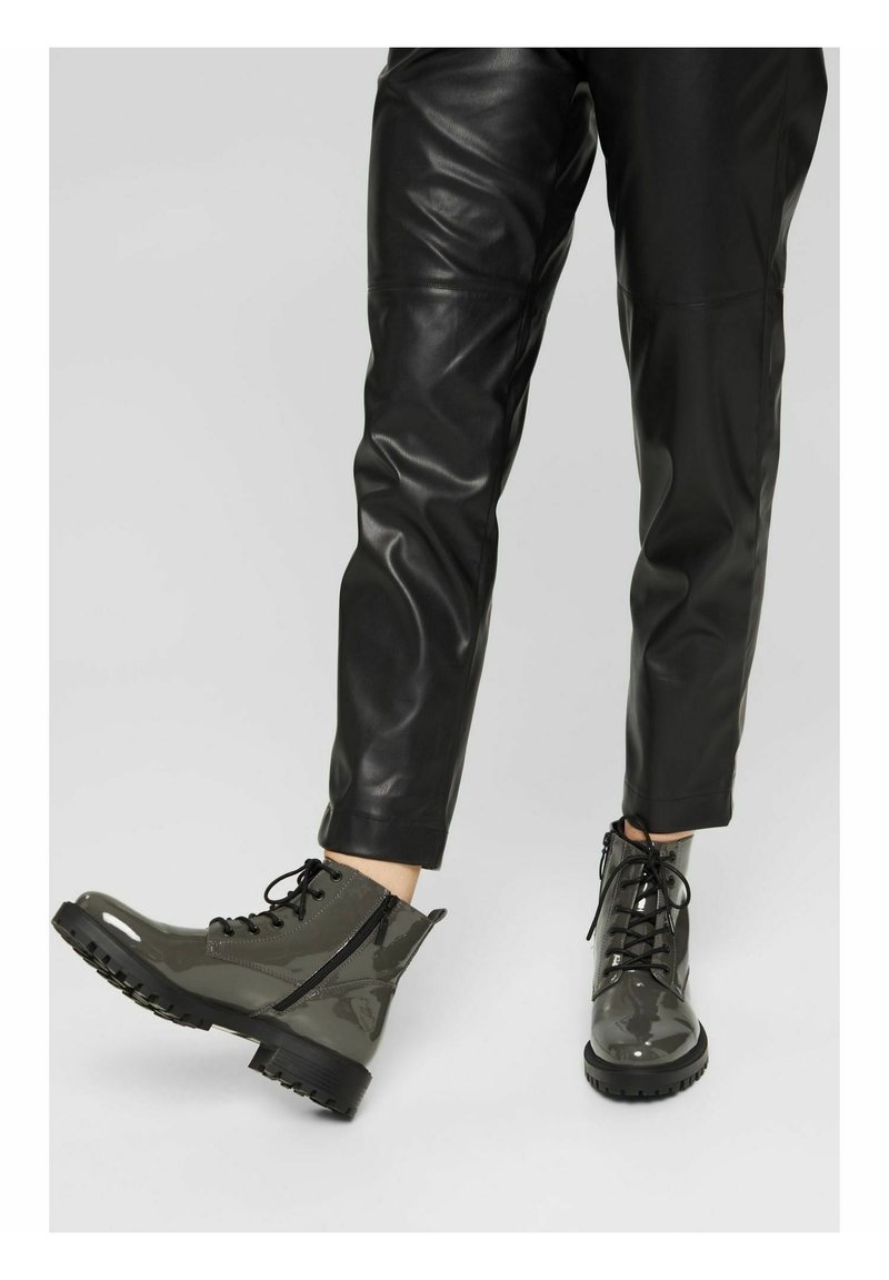 Personne portant un pantalon en cuir noir et des bottes de combat en cuir verni gris avec des semelles épaisses et des fermetures éclair latérales marchant sur un fond uni.