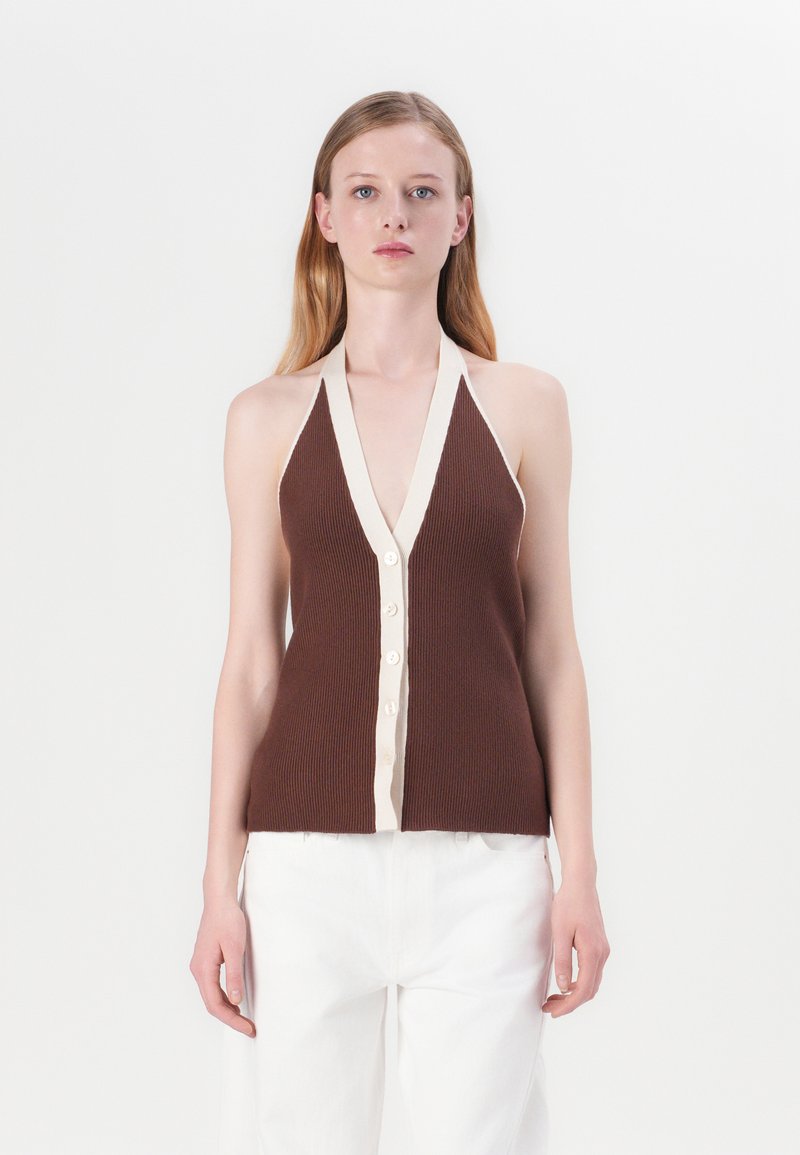 Hnedý pletený top s halter strihom a hlbokým V-výstrihom, béžové zapínanie na gombíky a ribbed textúra. V kombinácii s bielymi nohavicami pre kontrast.
