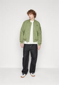 Giovane uomo con capelli ricci che indossa una giacca bomber verde, una maglietta bianca, jeans neri larghi e sneakers bianche con suole marroni.