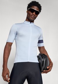 Tricou de ciclism albastru deschis cu fermoar pe jumătate, mâneci scurte, textură ribbed și o bandă bleumarin. De asemenea, sunt vizibile pantaloni scurți negri de ciclism și o cască neagră.