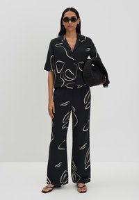 Ensemble deux pièces noir à motifs comprenant un haut à manches courtes et boutons ainsi qu'un pantalon à jambes larges, le tout orné de motifs abstraits crème. Les accessoires incluent un sac à main noir texturé.