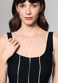 Srebrny naszyjnik z przezroczystym kamieniem w kształcie gruszki, noszony przez osobę w czarnej sukience w białe paski. Kolaż cery jest jasny.