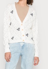 Vit stickad cardigan med diamantmönster, öppen fram och träknappar. Har marinblå blommönsterbroderi på de nedre sidorna.