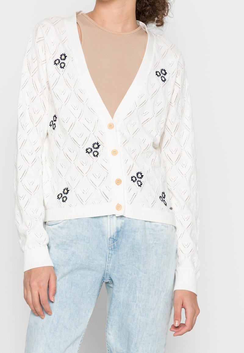 Vit stickad cardigan med diamantmönster, öppen fram och träknappar. Har marinblå blommönsterbroderi på de nedre sidorna.