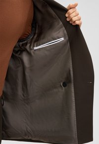 Bruin colbert met een patroon voering, sluiting met één knoop en een klein zakje met witte bies voor extra functionaliteit.