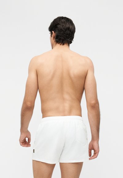 Άνδρας με σκούρα μαλλιά, χωρίς πουκάμισο, φορώντας λευκές βερμούδες, στέκεται με την πλάτη γυρισμένη σε απλό φωτεινό φόντο.