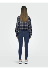 Camicia a quadri navy e crema, jeans blu aderenti e sneakers di colore chiaro. I lunghi capelli lisci scendono sulle spalle.