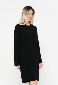Abito in maglia nera con maniche lunghe, scollatura rotonda e vestibilità ampia. Tessuto morbido, design fino al ginocchio e senza motivi o accessori visibili.