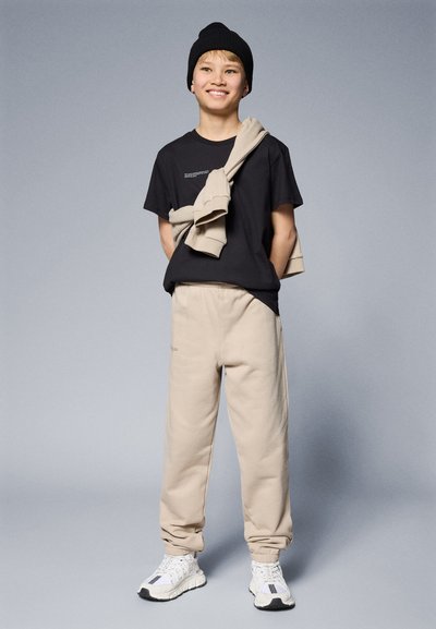Camiseta negra con texto impreso, pantalones de chándal beiges, zapatillas claras y un gorro negro, con un suéter colgado sobre los hombros.