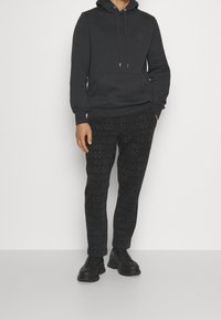 Haut à capuche gris foncé avec cordons de serrage et poche frontale, associé à un pantalon noir avec un motif logo répété, et des chaussures noires à lacets.