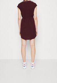 Burgundy kortärmad klänning med bälte i midjan, rund halsringning och en något böjd fåll, i kombination med vita sneakers med röda och blå detaljer.