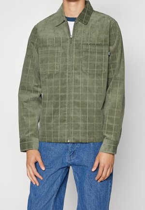 Veste en velours côtelé vert avec un motif à carreaux, fermeture éclair sur le devant, deux poches poitrine et manches longues. Assortie avec un jean en denim bleu.