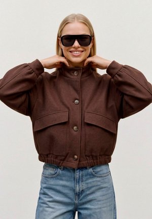 Une femme souriante portant de grandes lunettes de soleil noires, une veste marron boutonnée et un jean bleu ajuste son col relevé sur un fond uni.