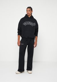 Μαύρη κουκούλα με γραφικό "ΔΕΝ ΥΠΑΡΧΕΙ ΣΧΕΔΙΟ Β", καγκουρό τσέπη και πλεκτές μανσέτες. Συνδυασμένο με ταιριαστά μαύρα παντελόνια φόρμας.