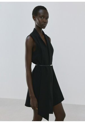 Mujer de pie con un vestido negro sin mangas, escote en V y cinturón delgado, sobre un fondo liso de color gris claro.