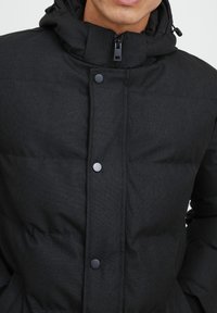 Sort polstret jakke med høj krave, frontlynlås og trykknapper. Stoffet har et struktureret mønster og en mat finish.