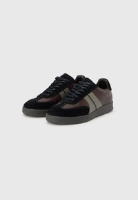 Black and brown suede sneakers feature a low-top design, gray accent stripes, and textured rubber soles with a slightly raised heel.