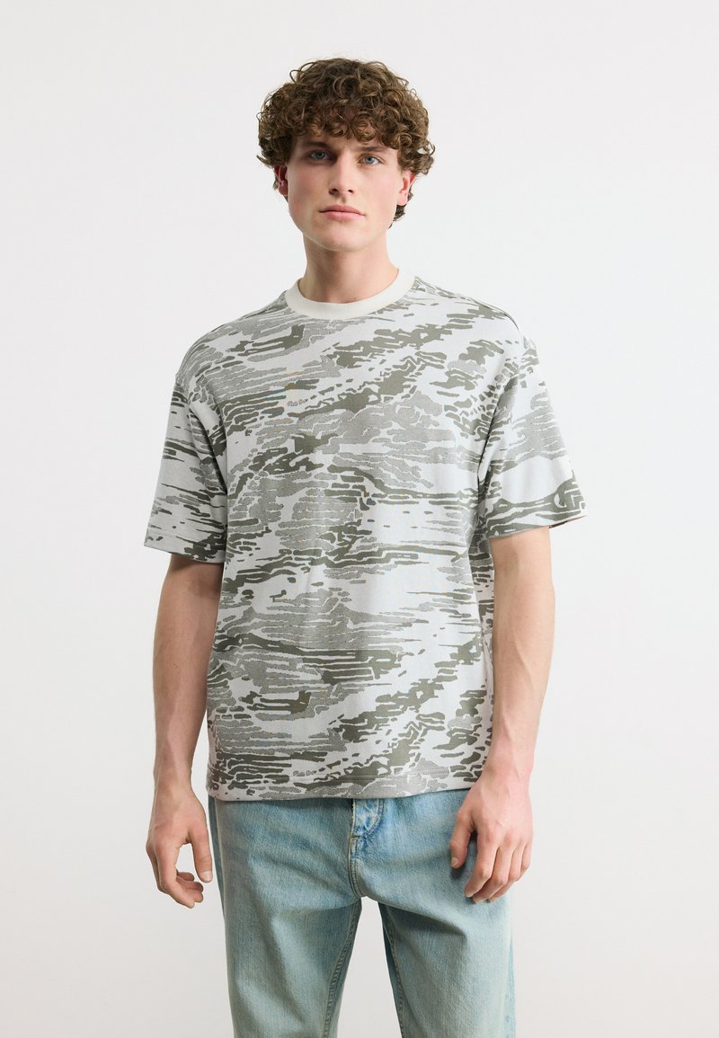 Νεαρός άνδρας με σγουρά μαλλιά που φοράει γκρι και λευκό πουκάμισο με κοντά μανίκια και αφηρημένο σχέδιο, καθώς και ανοιχτόχρωμα μπλε τζιν, μπροστά σε απλό φόντο.