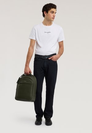 Giovane uomo che indossa una t-shirt bianca Trussardi e jeans neri, che tiene uno zaino in pelle verde oliva con una mano, guardando di lato.