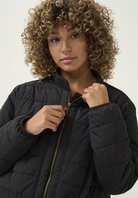 Chaqueta acolchada negra con cuello alto, cremallera dorada y puños elásticos. La tela parece suave y ligeramente brillante.