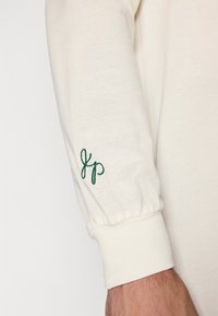 Langermet topp i off-white bomull med ribbede mansjetter og en grønn brodert logo på venstre arm. Strukturert stoff med subtilt mønster.