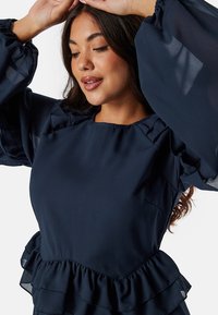 Marineblauw peplumtop met ruches, lange doorschijnende mouwen en een gladde textuur. Heeft een ronde halslijn en een getailleerde taille.