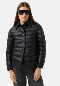 Schwarze Steppjacke mit glänzendem Finish, mit Kragen, Reißverschlusstaschen und silbernen Druckknöpfen. Gestylt mit übergroßen Sonnenbrillen.