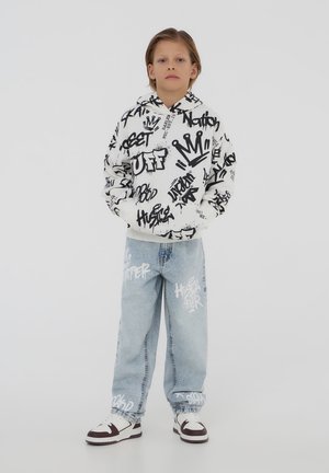Felpa bianca con motivi graffiti neri, abbinata a jeans di denim azzurro chiaro con design di testo bianco e sneaker in bianco e nero.
