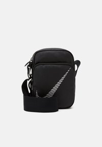 Černá nylonová taška přes rameno s nastavitelným popruhem, s velkým šedým logem Nike swoosh a zipovým uzavřením. Texturovaný povrch.