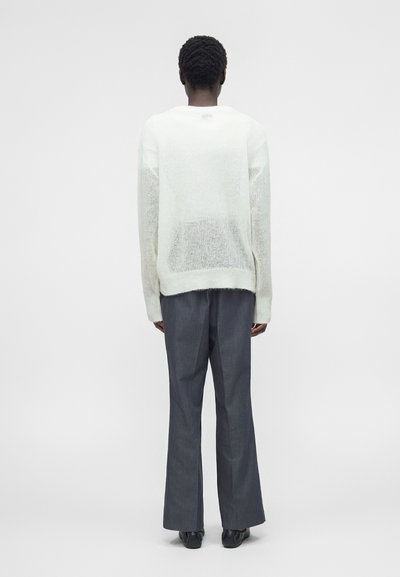 Άτομο με κοντά σγουρά μαλλιά που φοράει λευκό πλεκτό πουλόβερ και γκρι παντελόνια με φαρδιά μπατζάκια, στέκεται γυρισμένο με την πλάτη στον θεατή.