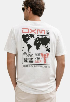 Uomo che indossa una t-shirt bianca con una mappa del mondo nera e icone grafiche rosse stampate sulla schiena, in piedi con le mani in tasca.