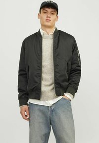 Sort bomberjakke i et glat materiale, med lynlås foran, sidelommer og ribbede manchetter, lagd over en lysegrå sweater.