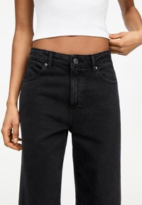 Femme portant un jean noir taille haute et un top court blanc, ajustant le haut d'une main.