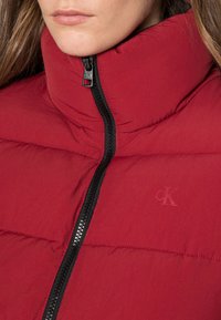Czerwona kurtka puchowa z wysokim kołnierzem, z czarnym zamkiem błyskawicznym i subtelnym logo. Materiał wygląda na lekki i pikowany dla lepszej izolacji.