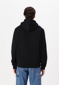 Personne avec les cheveux bruns courts portant un sweatshirt à capuche noir et un jean cargo en denim bleu, tourné le dos contre un fond uni.