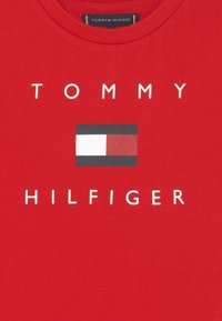 Camisa roja con el texto blanco "TOMMY HILFIGER" y un logotipo rectangular que presenta bloques de color blanco, rojo y negro, centrado debajo de "TOMMY".