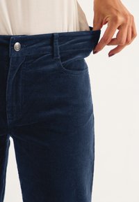 Navy corduroy pants with a high waist, front pockets, and a button closure; fabric has a textured, ribbed appearance.