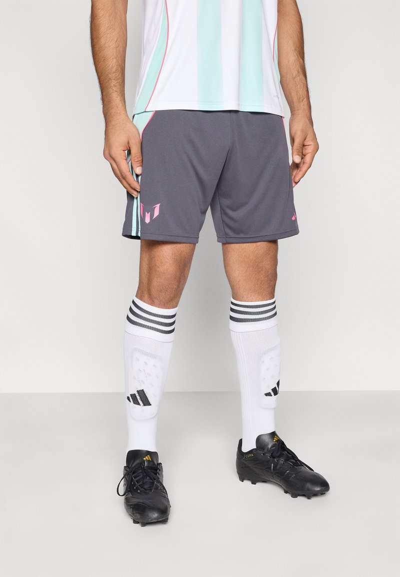 Γκρι αθλητικό σορτς με ροζ λεπτομέρεια λογότυπου, συνδυασμένο με λευκές κάλτσες με μαύρες ρίγες και μαύρα παπούτσια ποδοσφαίρου.