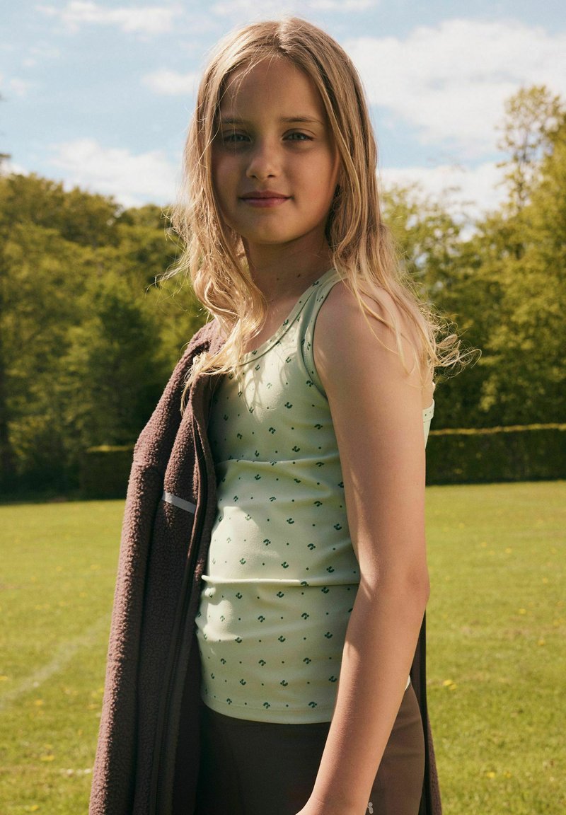 Jeune fille aux longs cheveux blonds portant un débardeur vert clair et tenant une veste sombre à l'extérieur par une journée ensoleillée avec des arbres en arrière-plan.