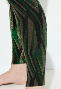 Leggings avec une base noire ornée de motifs en zigzag verts et dorés, fabriqués dans un matériau métallique texturé qui scintille.